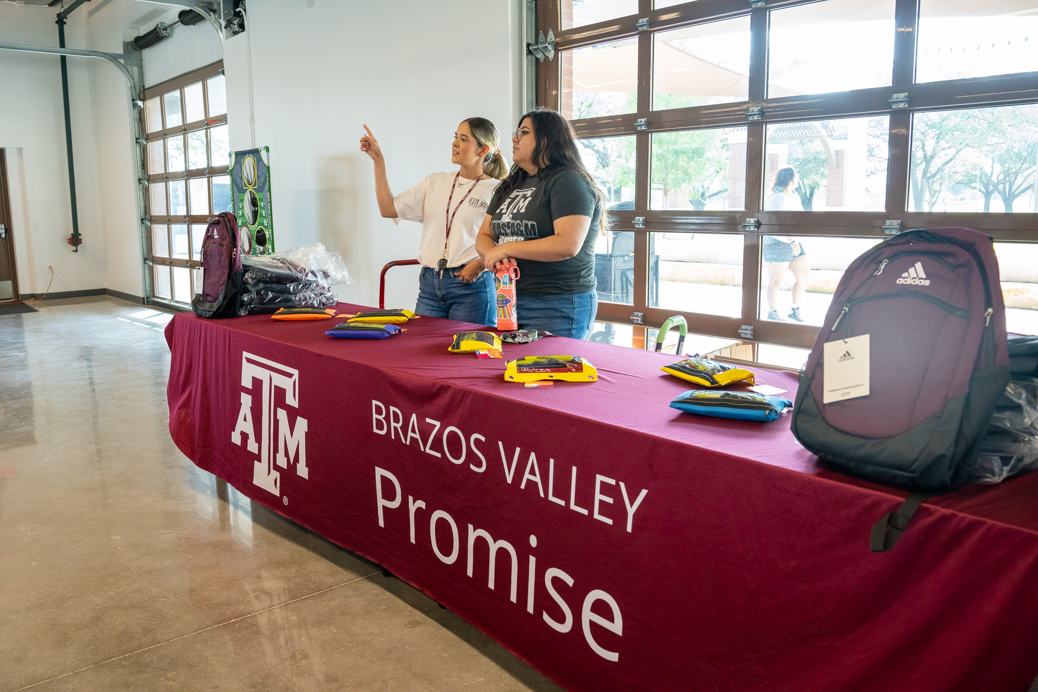 Two women stand behind a long table covered with a maroon tablecloth featuring the Texas A&M logo and the text "Brazos Valley Promise." One woman points toward something out of frame while the other looks on. The table is lined with maroon Adidas backpacks and small blue and yellow pouches. Large glass garage-style doors in the background look out onto a sunlit outdoor area.